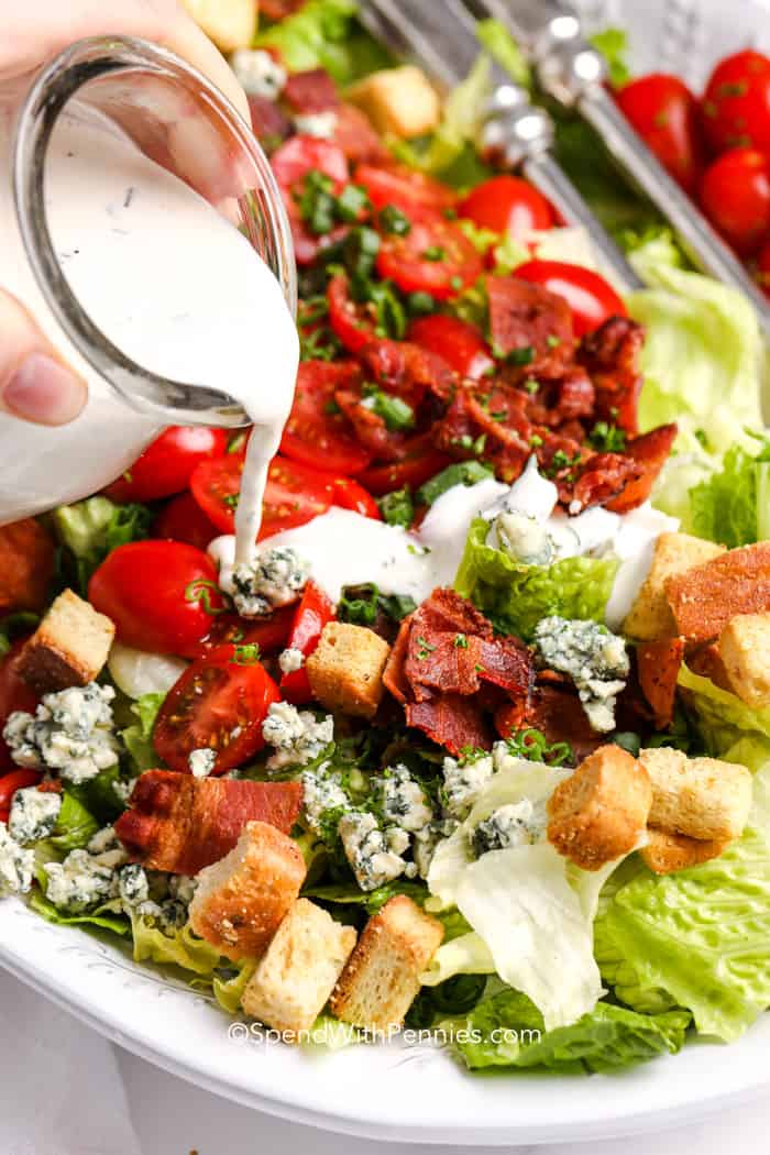 BLT salad blends fresh greens, juicy tomatoes, smoky bacon, and crumbled cheese into a salad that eats like a meal. The dressing is creamy with a slight tang, made with sour cream, mayo, and dill for a burst of flavor. It’s great for serving at BBQs, picnics, or family dinners. This easy-to-make salad is a new favorite in any meal rotation. #spendwithpennies pouring a creamy herbed dressing over a blt salad