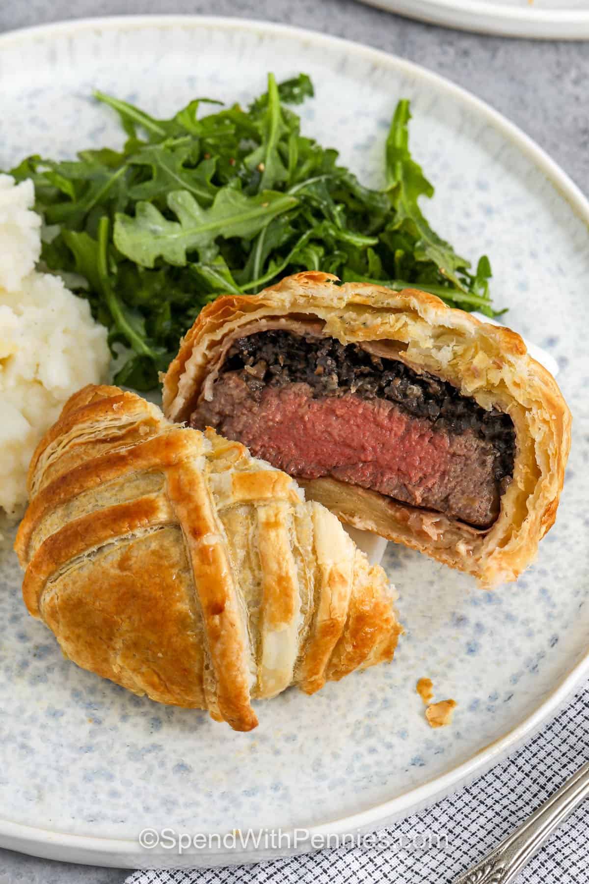 A flaky crust, juicy filet mignon, and garlicky mushrooms make this Beef Wellington recipe a dinner to remember. It’s one of those showstopper meals that turns out beautiful every time with a few easy steps. The prosciutto adds a salty bite that pairs perfectly with the rich filling, and that Dijon mustard gives it the perfect little zing. It's special enough for guests, but easy enough for anytime! #spendwithpennies Beef Wellington sliced in half on a plate