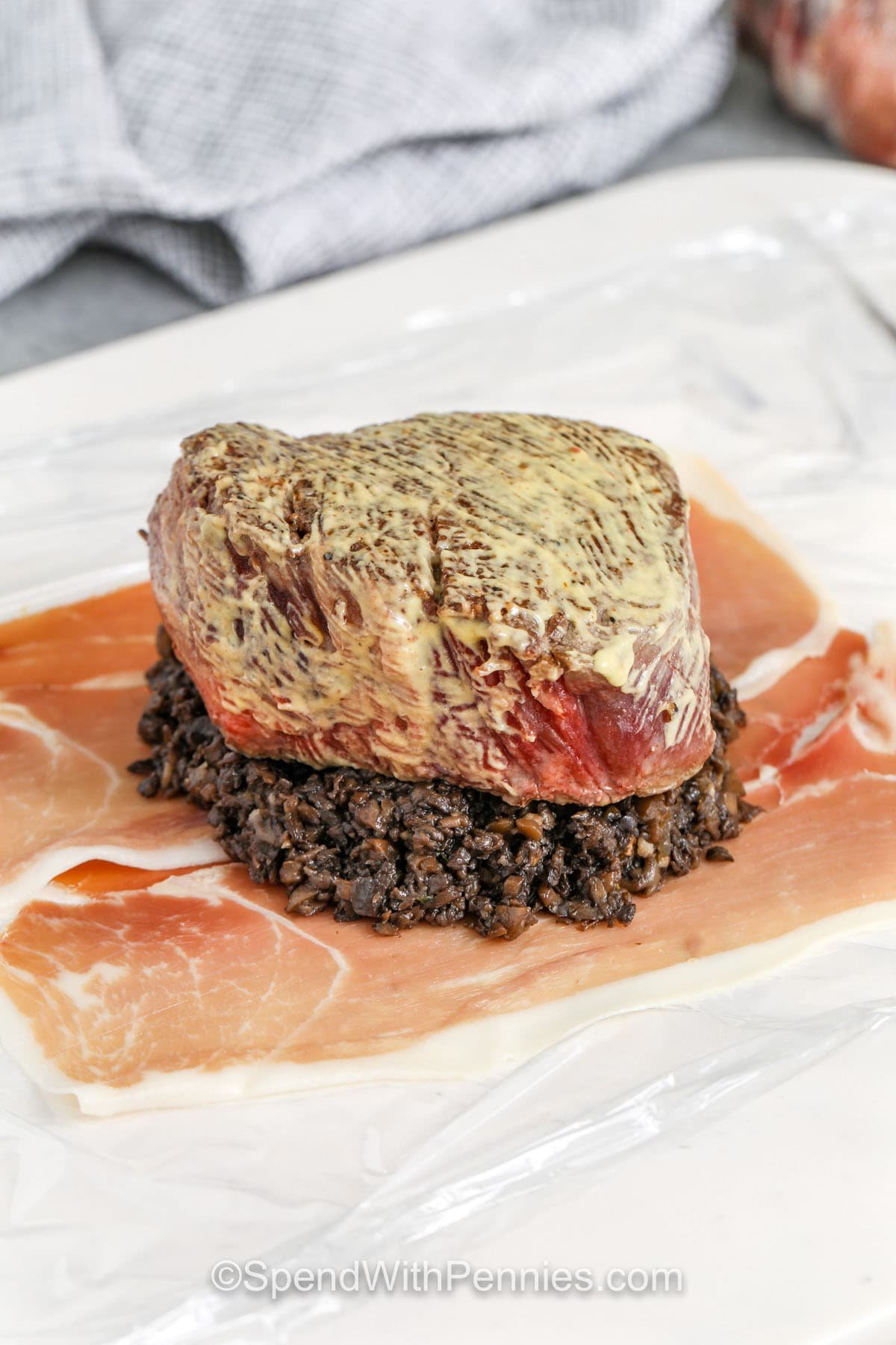 Looking for something a little extra special, this Beef Wellington recipe is just the thing. A buttery puff pastry shell, perfectly seared steak, and a savory mushroom filling make every bite melt-in-your-mouth delicious. It’s elegant enough to serve for a holiday or celebration but simple enough to enjoy any time. You’ll love how the flavors all come together in one beautiful little package! #spendwithpennies adding beef to mushroom mixture on prosciutto to make beef wellington