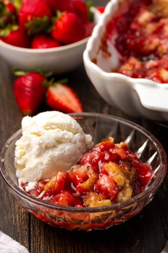 A serving of strawberry cobbler in a bowl topped with vanilla ice cream, with the rest of the cobbler in a baking dish in the background.