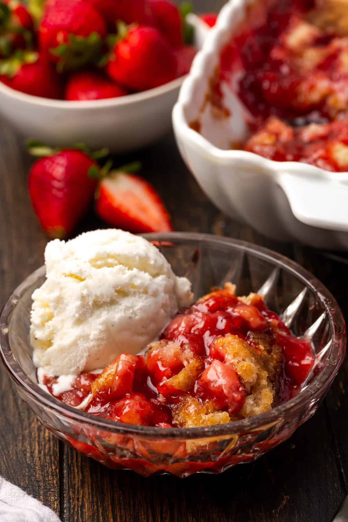 A serving of strawberry cobbler in a bowl topped with vanilla ice cream, with the rest of the cobbler in a baking dish in the background.