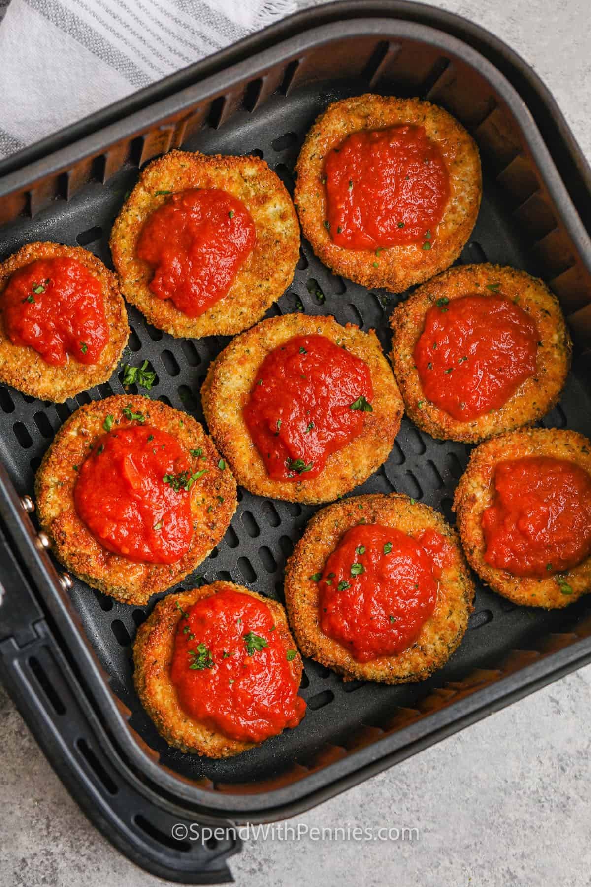 Get ready for easy air fryer eggplant parmesan to become your favorite way to make this delicious meal. Slices of eggplant are breaded in a savory breadcrumb mixture containing parmesan, herbs, and garlic powder, then they’re air fried until crispy. Top the slices with marinara sauce and mozzarella, and cook until melted. Try swapping thin chicken cutlets for the eggplant. For a keto version, leave out the bread crumbs. #spendwithpennies adding marinara sauce to Air Fryer Eggplant Parmesan in the fryer