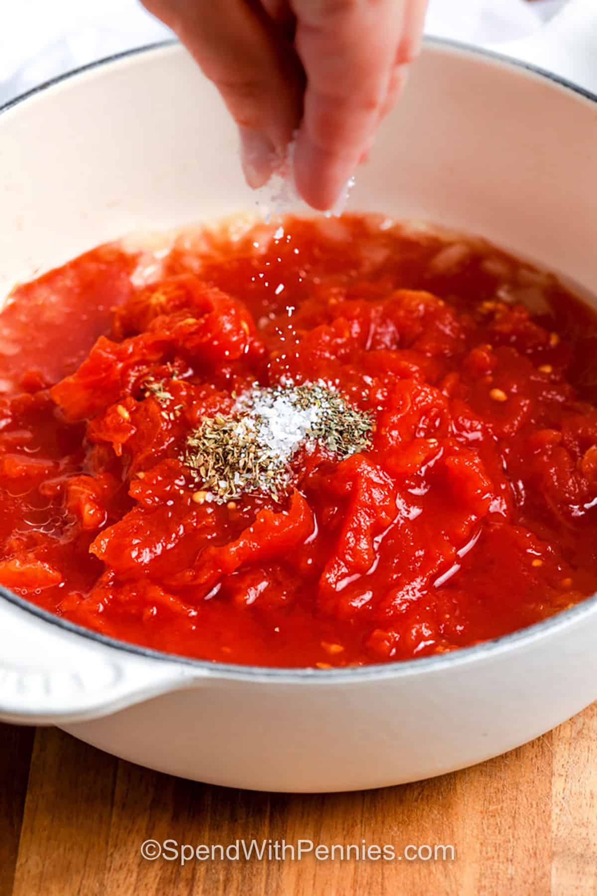 Tomato sauce from scratch is simple, satisfying, and so much tastier than anything from a jar. Sautéed onions and garlic lay the flavor foundation, while hand-crushed canned tomatoes, basil, and parsley add richness and herby brightness. This sauce is great to make ahead and freezes beautifully for future meals. A true recipe essential for pasta or spaghetti night! #spendwithpennies adding seasoning to tomatoes to make Tomato Sauce